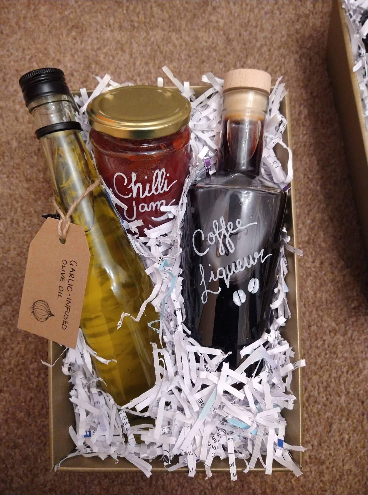 A homemade hamper with a bottle of garlic olive oil, chilli jam, and coffee liqueur surrounded by shredded paper.