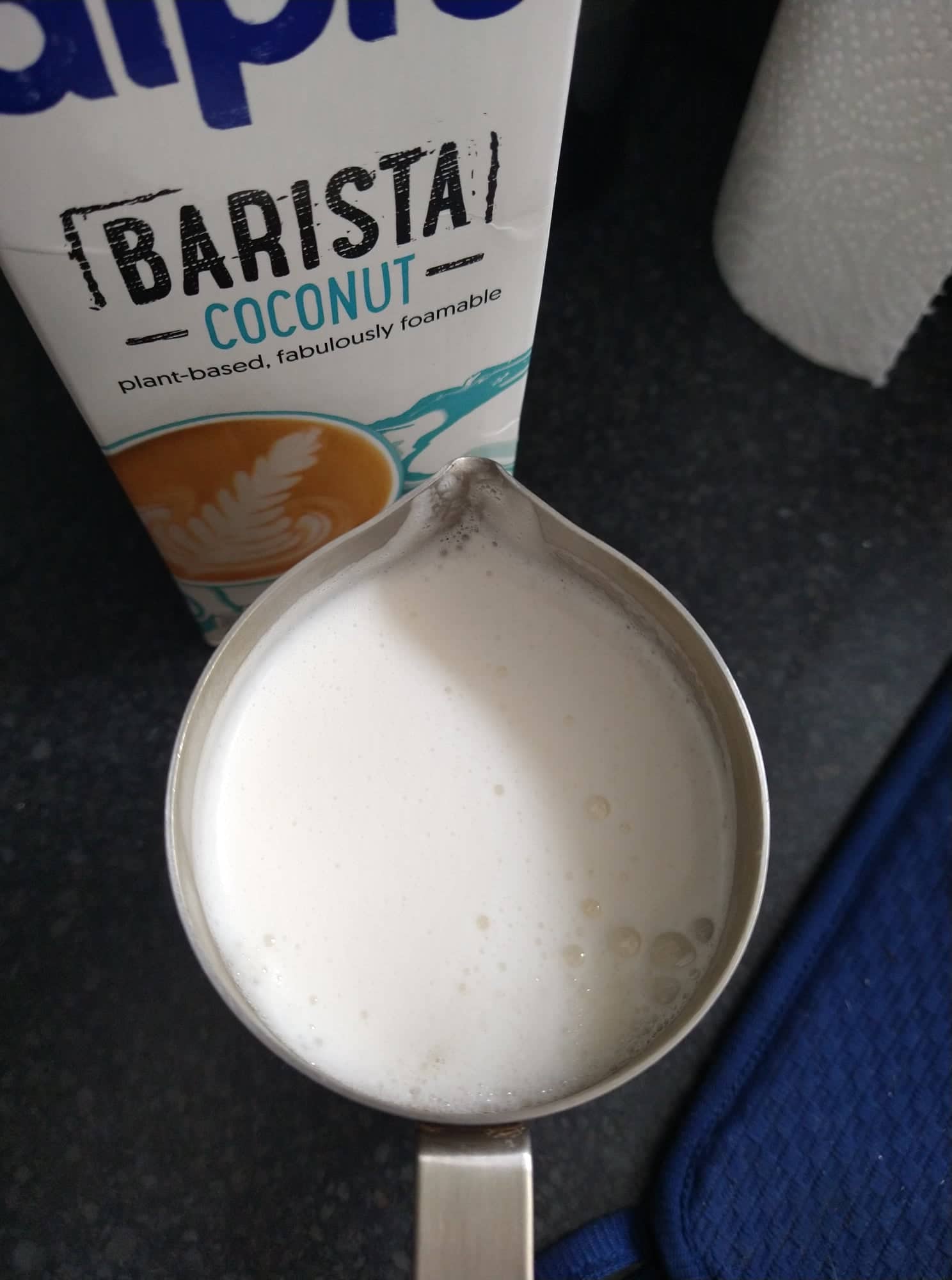Creamy and frothy coconut milk in the frothing jug, beside a carton of Alpro barista coconut.