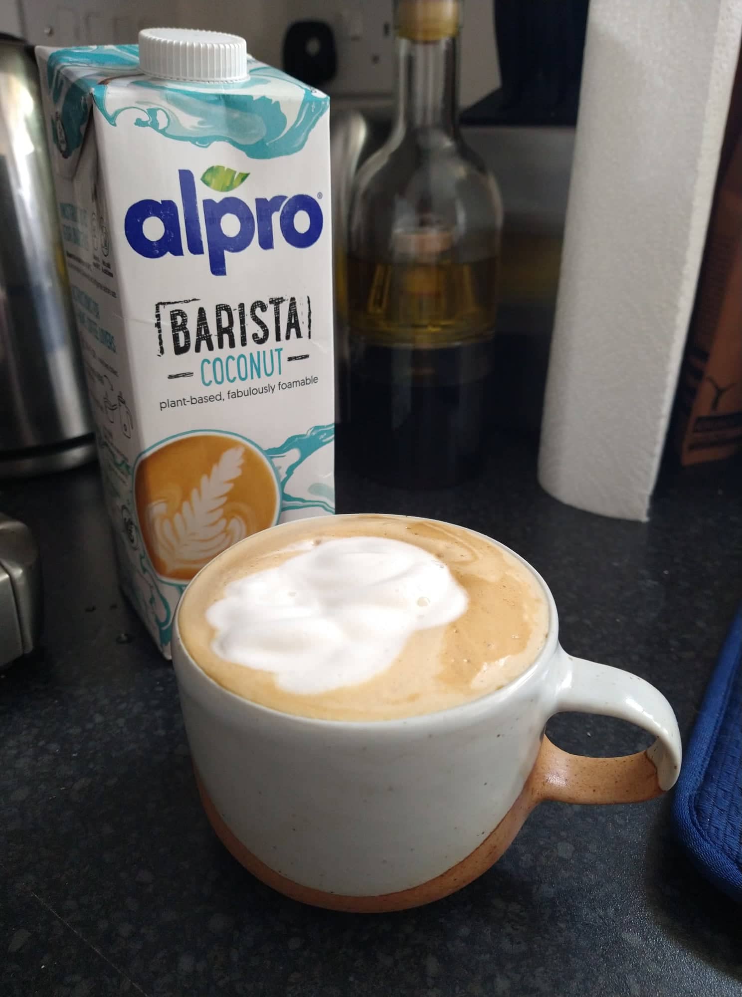 A full cup of coffee with too much white and brown foam that's come out in a blob, beside a carton of Alpro barista coconut.