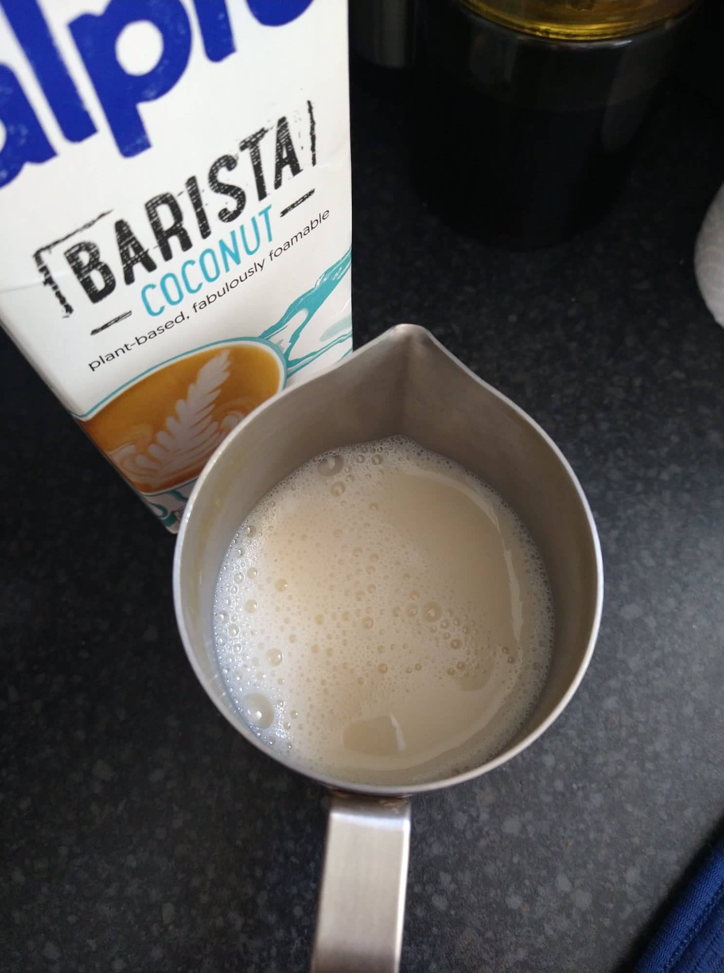 Beige coconut milk in the frothing jug, pre-froth, beside a carton of Alpro barista coconut.
