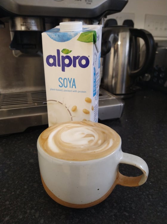 A foamy coffee made with soya milk and the white carton of Alpro soya milk, in front of our Sage coffee machine.