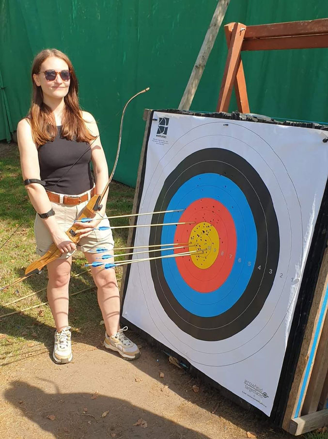 Me stood in the sun at Sherwood Forest, holding a bow beside a target full of arrows.