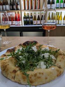 A freshly made pizza covered in rocket, and two glasses of wine at Okende in West Bridgford. Behind the table is a wall of shelves filled with various wine bottles.