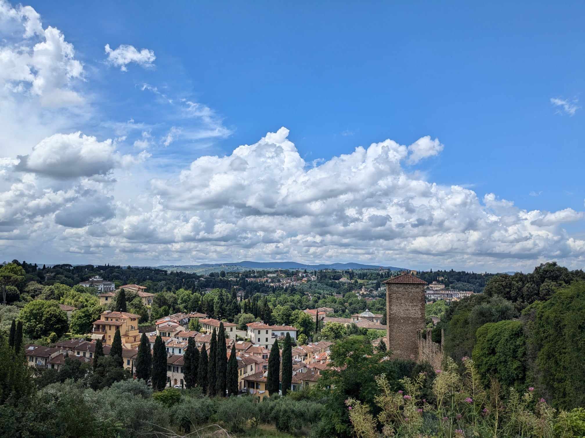 A view over sunny Tuscan countryside, with tall cypress trees, and small white buildings with red roofs.