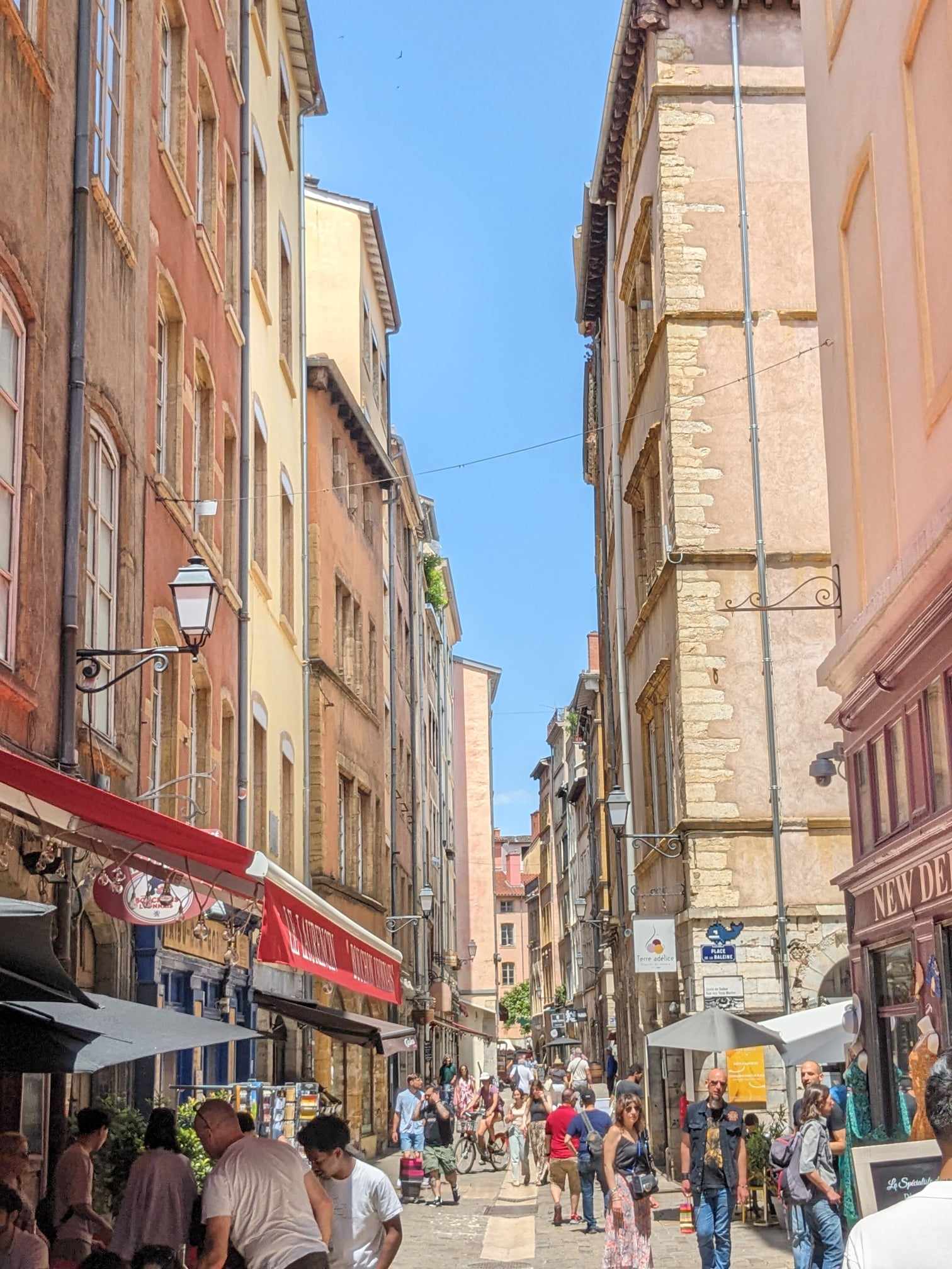 A sunny street in Lyon, with tall red buildings, and tourists looking into shops under awnings.