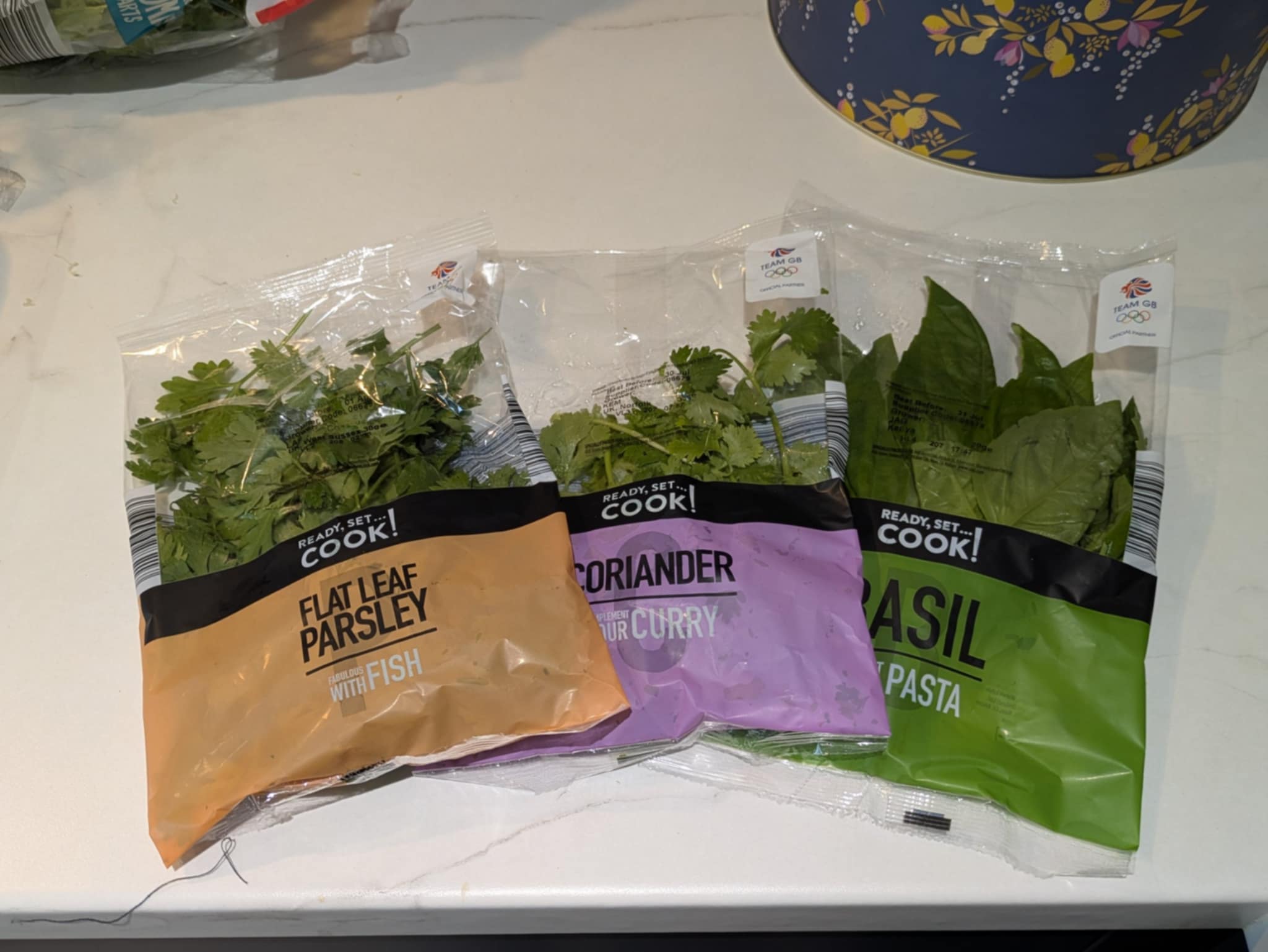 Three bags of herbs on a counter: parsley, coriander, and basil.